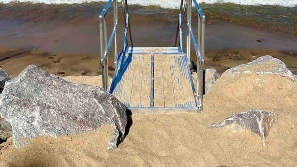 Una playa con una escalera de madera que baja hacia el agua, rodeada de grandes rocas.