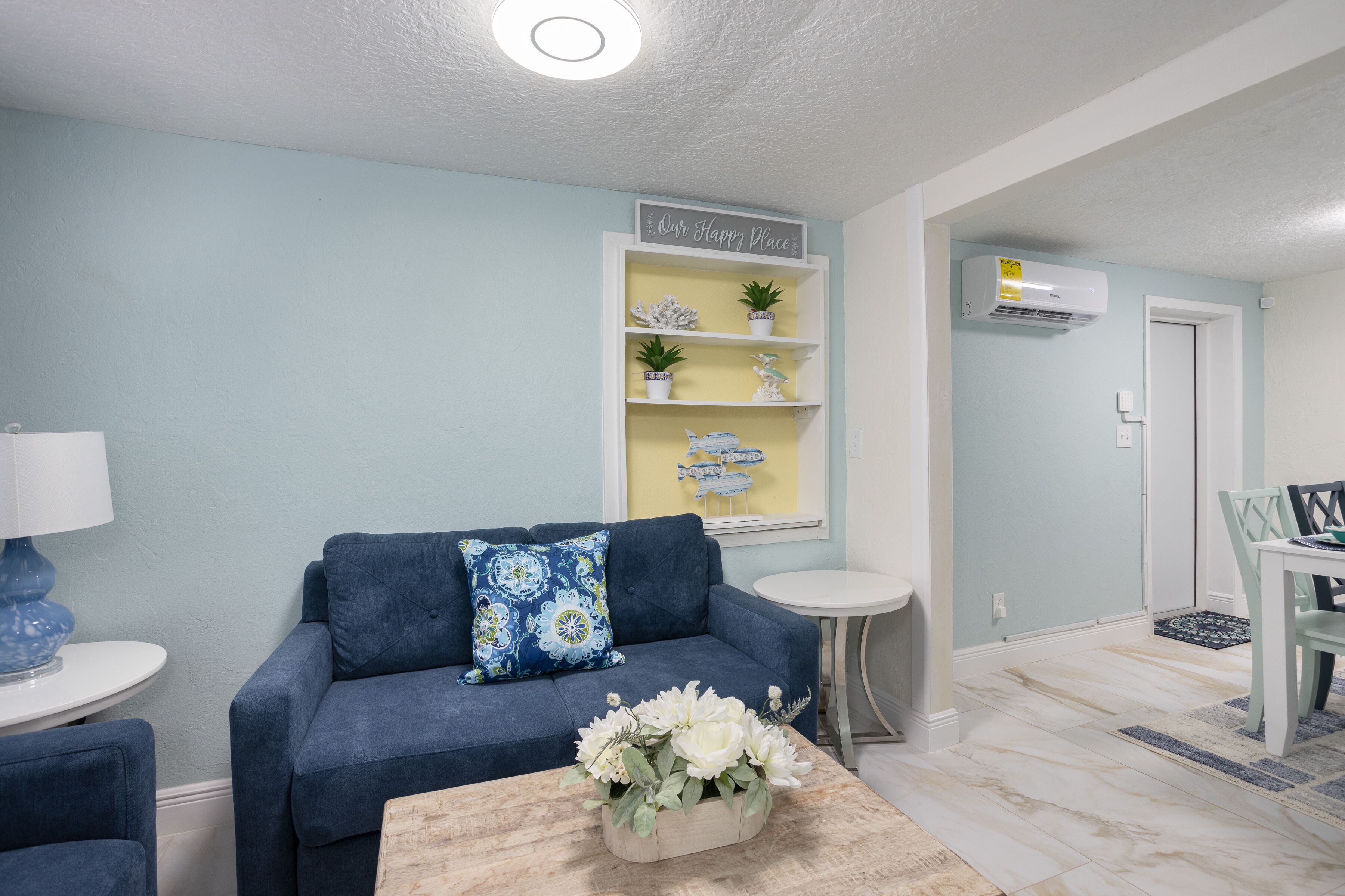 A living room with a blue sofa, a small round table, and a coffee table with flowers. There is a shelf with decorative items and a sign that says "Our Happy Place".