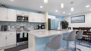 A modern kitchen with white cabinets, stainless steel appliances, a blue backsplash, and a bar area with two bar stools. - Beach Getaway on Cocoa Beach (Cocoa Beach)