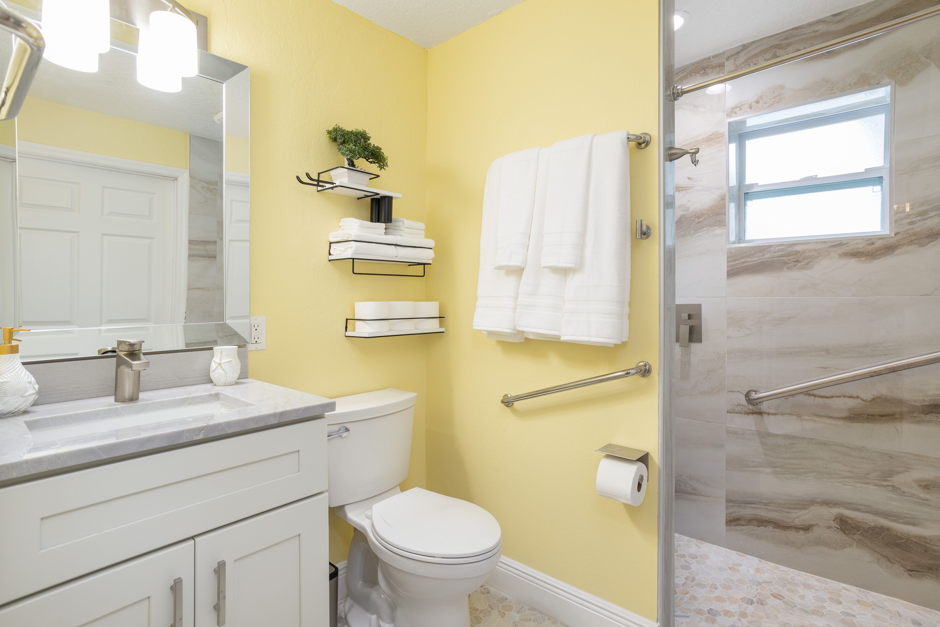 A bathroom with a yellow wall, white fixtures, a glass shower enclosure, and a small window.