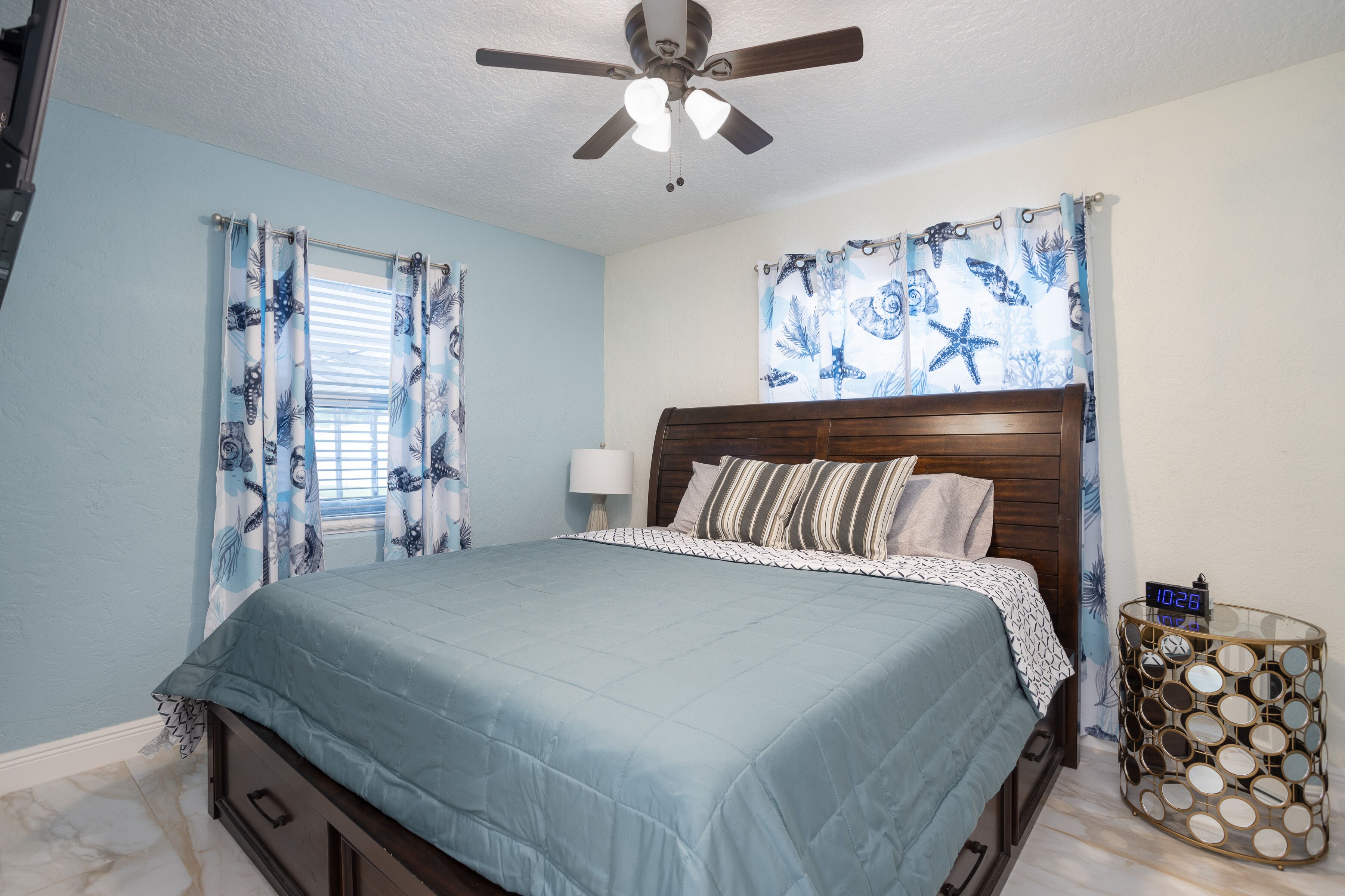 A bedroom with a bed, a ceiling fan, a nightstand with a clock, and curtains with a marine theme.