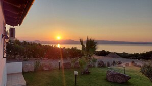 A sunset view from a house with a garden in front, overlooking a calm body of water and distant mountains.