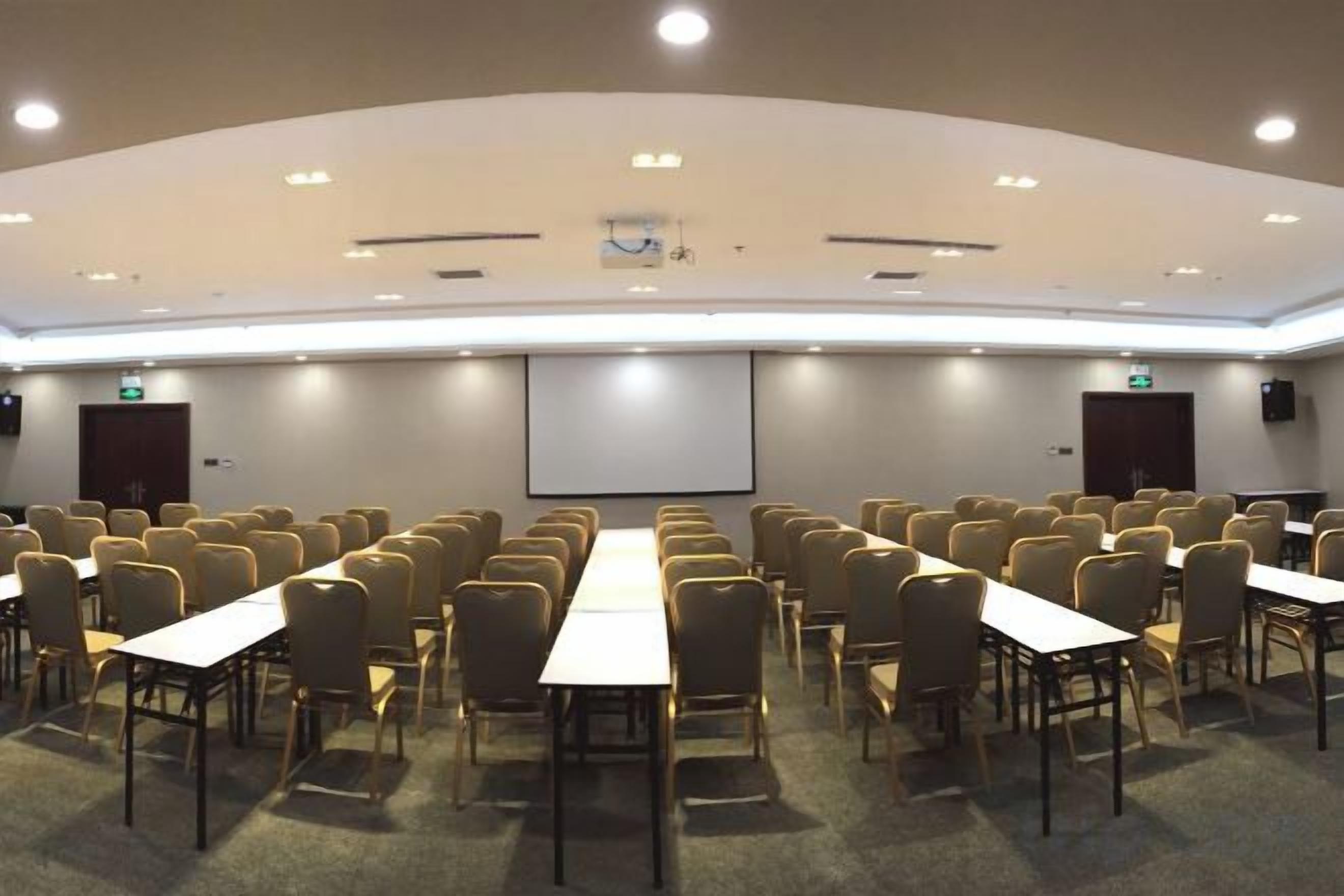 Une salle de conférence avec des rangées de chaises et de tables, un écran de projection et un tableau blanc.