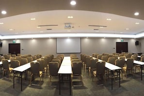 Una sala de conferències amb files de cadires i taules, una pantalla de projector i una pissarra.