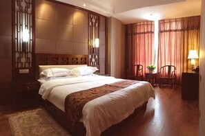 A hotel room with a large bed, a wooden headboard, a nightstand, two chairs, a small table, and a lamp. - Manas County Hotel (Manas County)