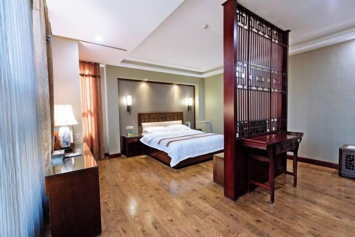 A hotel room with a wooden floor, a bed with white linens, a nightstand, a desk with a lamp, and a traditional wooden partition.