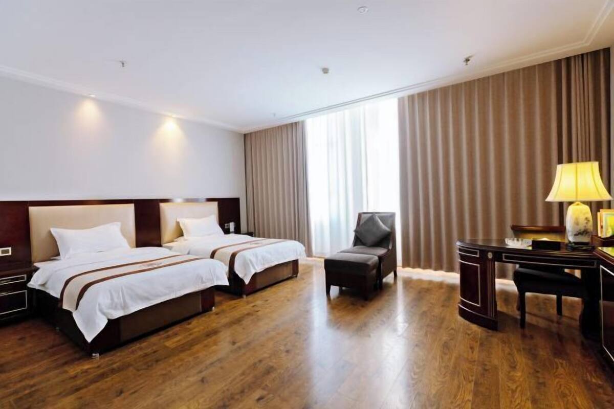 A hotel room with two beds, a desk, a chair, and a lamp.