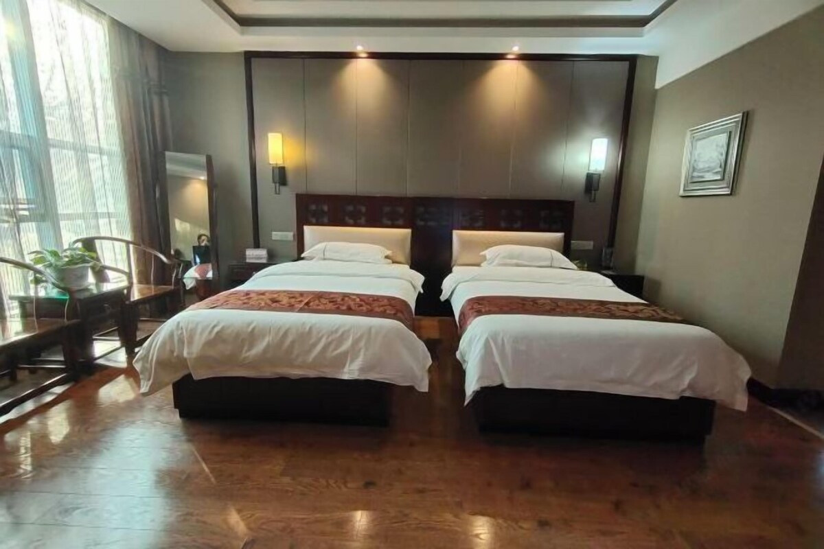 A hotel room with two beds, wooden flooring, a large window with curtains, and a small table with a plant.