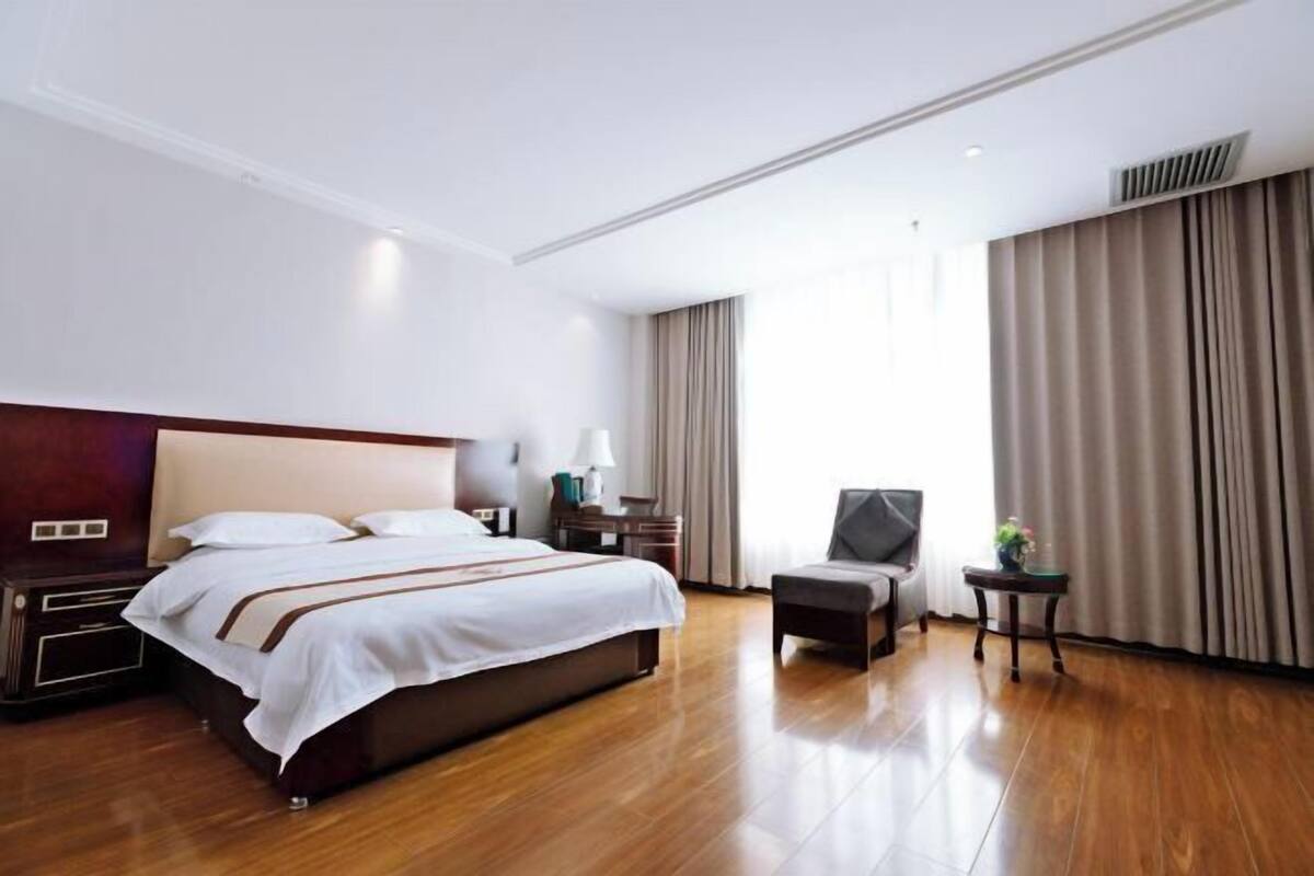 A hotel room with a large bed, bedside tables, a chair, a small table with a lamp, and a window with curtains.