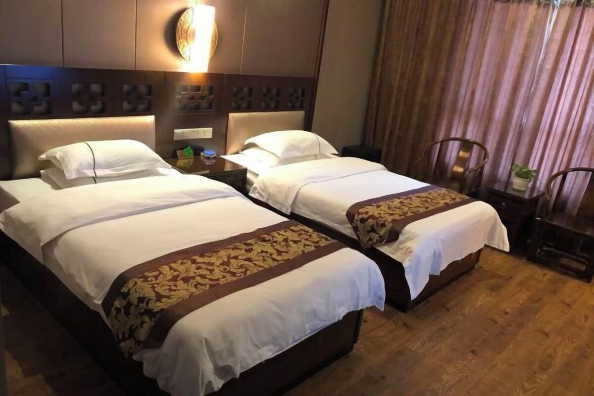 A hotel room with two beds, each with white linens and a brown decorative border. There is a wall-mounted light fixture above the beds and a curtain to the right.