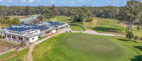 An aerial view of a golf course with a large building featuring solar panels, surrounded by lush greenery and trees.