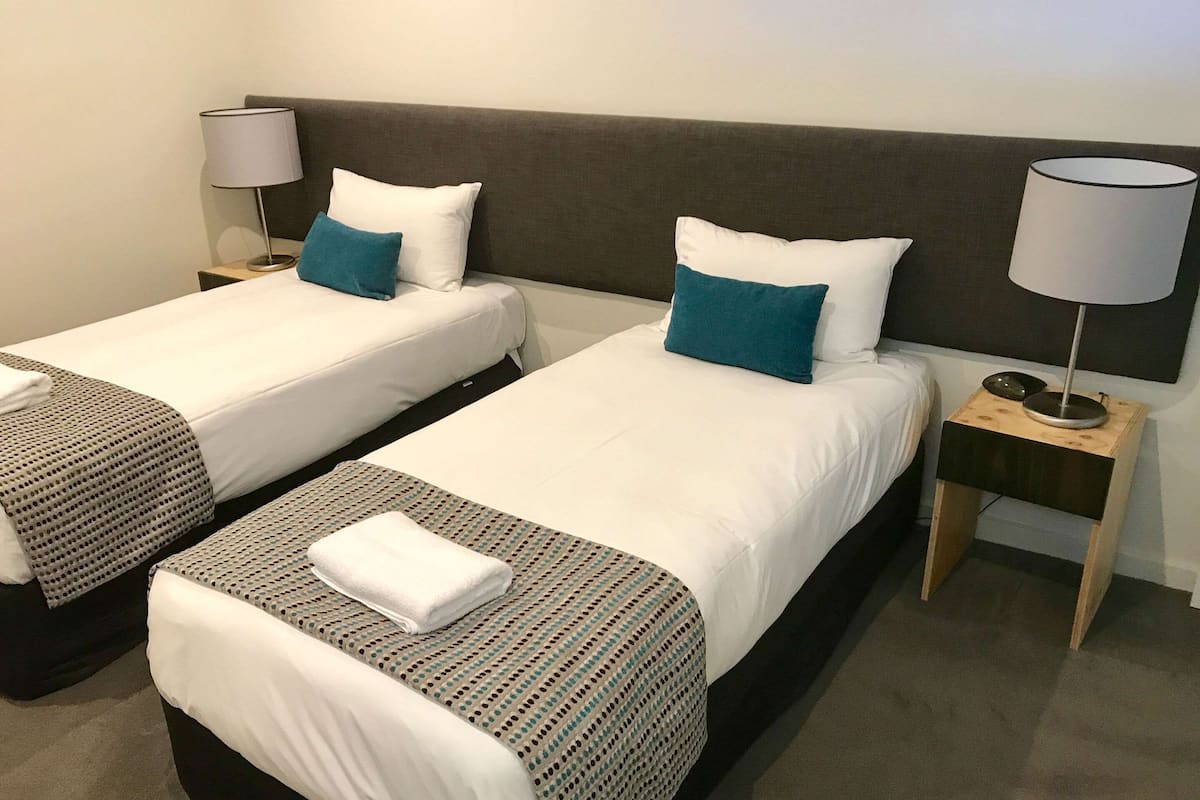 A hotel room with two single beds, each with a pillow, a bedside table with a lamp, and a blanket.