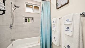 A bathroom with a bathtub, a shower, and a window with a frosted glass door. There are towels hanging on the wall and a framed picture above the window.