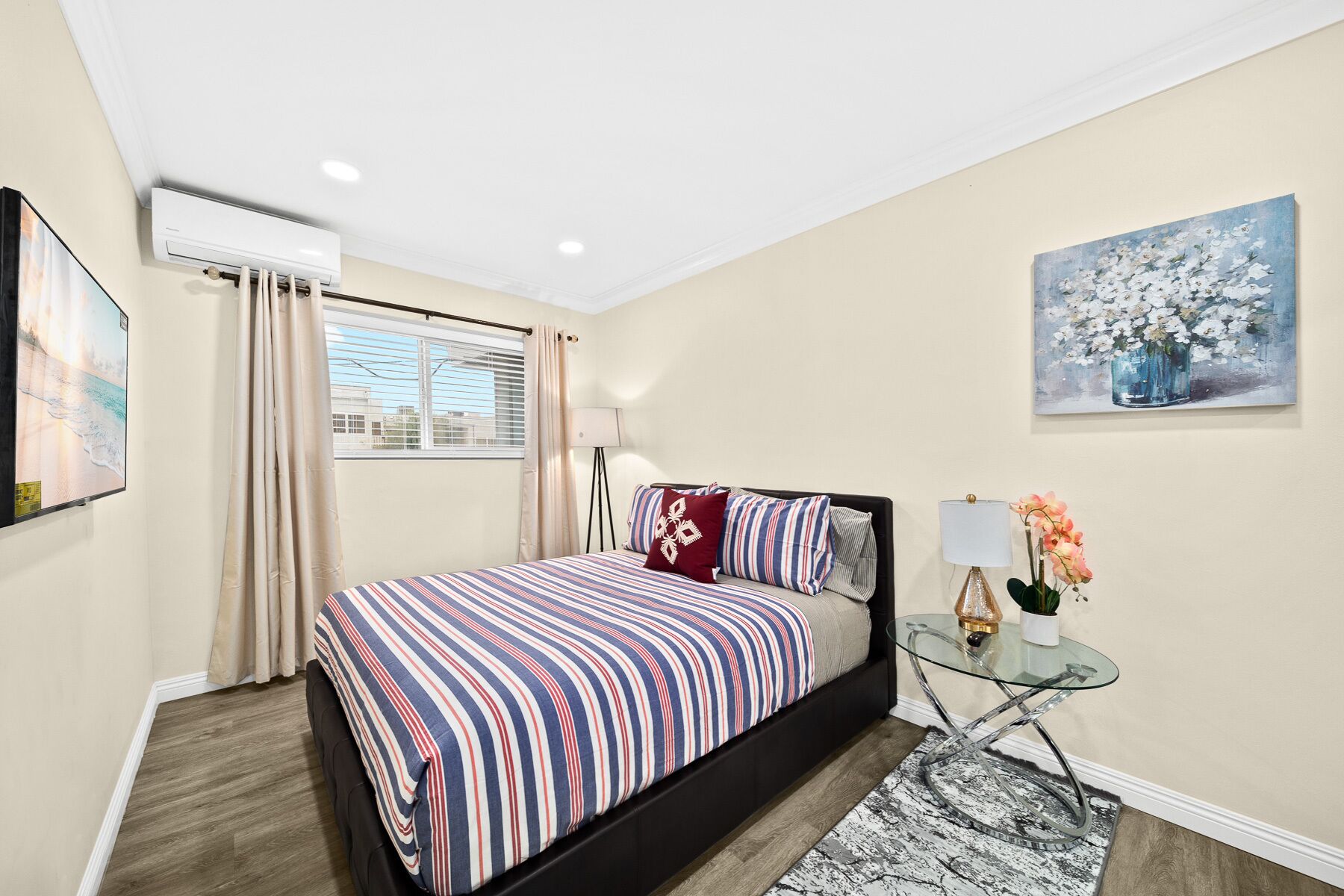 A bedroom with a bed, a bedside table with a lamp, a painting on the wall, and a window with curtains.