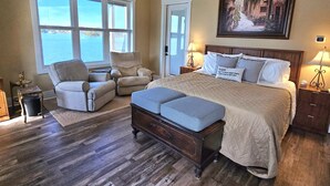 Een slaapkamer met een groot bed, twee fauteuils, een houten vloer, een raam met uitzicht en een schilderij aan de muur.