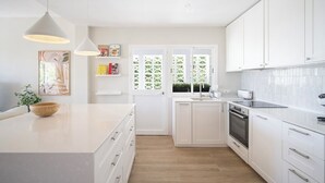Une cuisine moderne avec des meubles blancs, un îlot central, des luminaires suspendus et une vue sur l’extérieur à travers une fenêtre ornée d’un treillage vert.