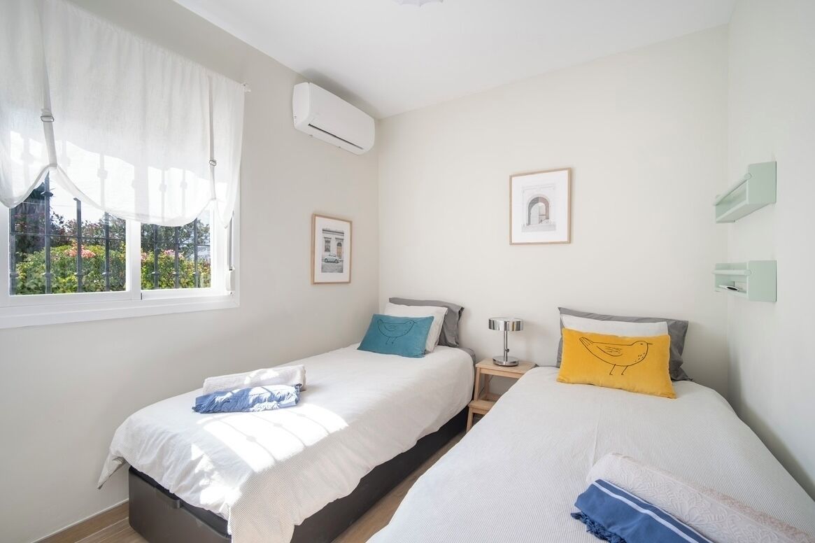 A hotel room with two beds, a window with curtains, an air conditioning unit, framed pictures on the wall, and a small bedside table with a lamp.