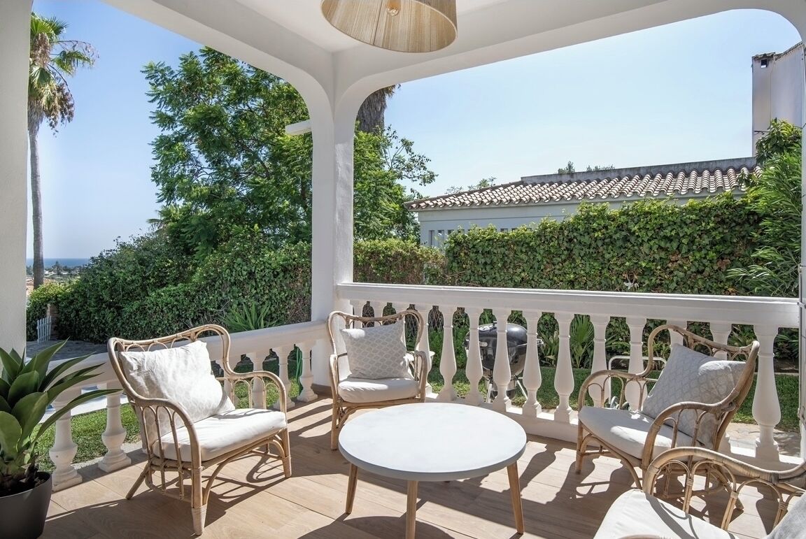 A covered outdoor seating area with wicker furniture and a round table, overlooking a garden and a building with a tiled roof.