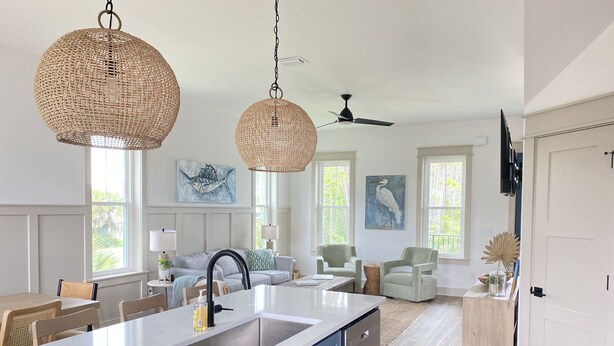 Una cucina moderna con una grande isola, mobili bianchi e un piano di lavoro blu. Sono presenti lampade pendenti in vimini, una ventola a soffitto e opere d'arte alle pareti.