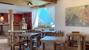 Uma área de jantar rústica com mesas e cadeiras de madeira, um ventilador de teto e uma grande janela com cortinas.