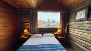 Quarto de cabana de tronco com cama, duas luminárias de cabeceira, um telefone e uma janela com cortinas.