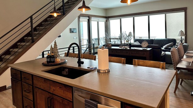 A modern kitchen with a central island, wooden cabinets, and a stainless steel sink. There is a staircase with a metal railing leading to an upper level. The room has large windows, a dining area with a table and chairs, and a living room with a sofa and a coffee table. - New, modern mountain retreat for several families 8 min from ski resort. (Fraser)