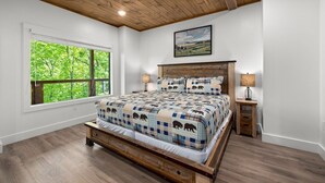 A bedroom with a wooden ceiling, a large window with a view of trees, a bed with a quilted blanket, and two bedside tables with lamps. - Modern, Cozy with Stunning Mountain View, Hot Tub, Arcade, Mins to the Smokies (Seiverville)