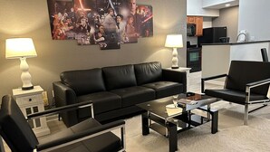 A modern living room with a black leather sofa, a glass coffee table, and a black armchair. There is a wall-mounted artwork depicting a movie scene. - Close to Disney and Universal! Family-sized condo with pool, balcony & game room (Orlando)