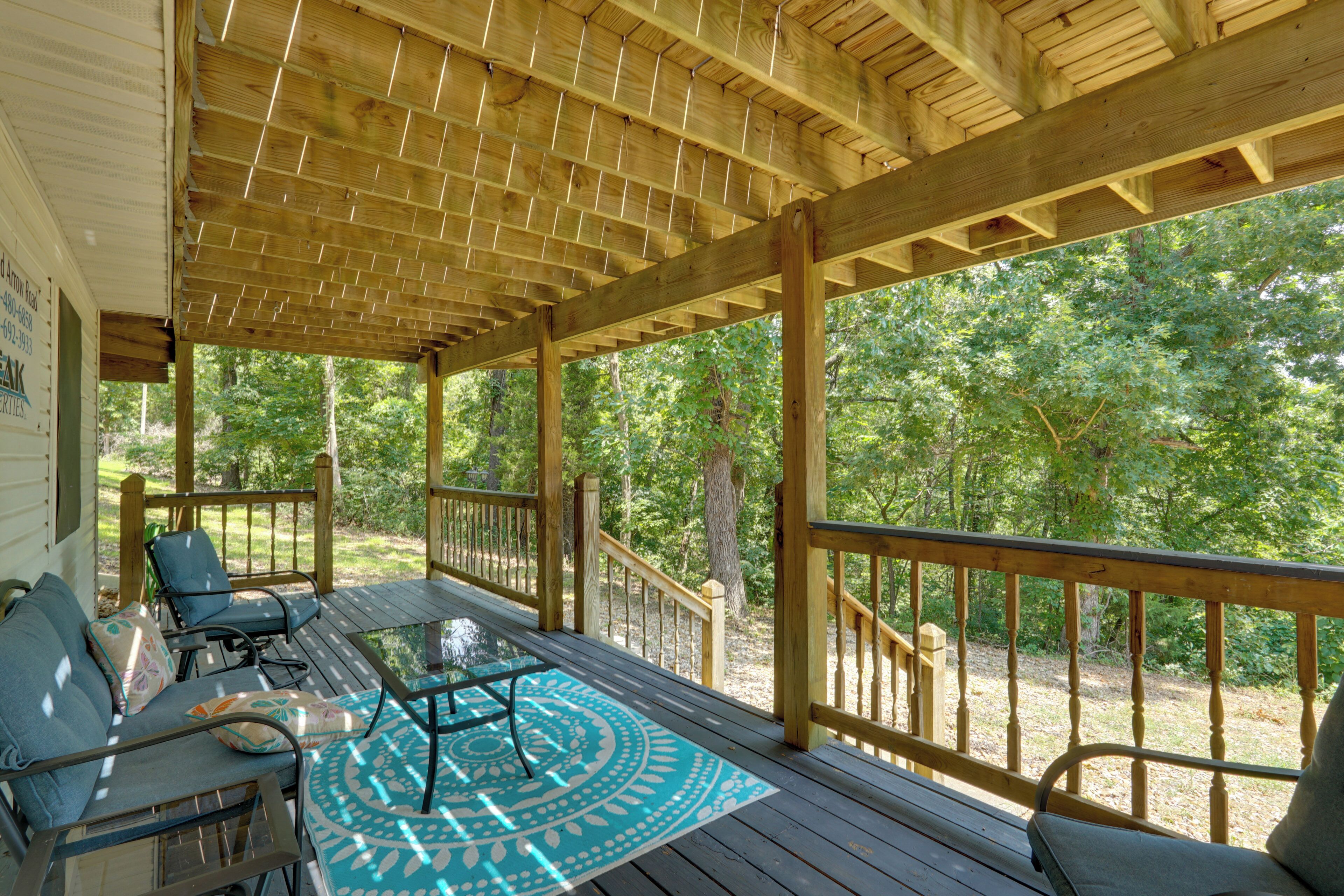 Une terrasse extérieure couverte, dotée de rampes en bois, d’une table en verre et d’un tapis d’extérieur à motifs bleus.