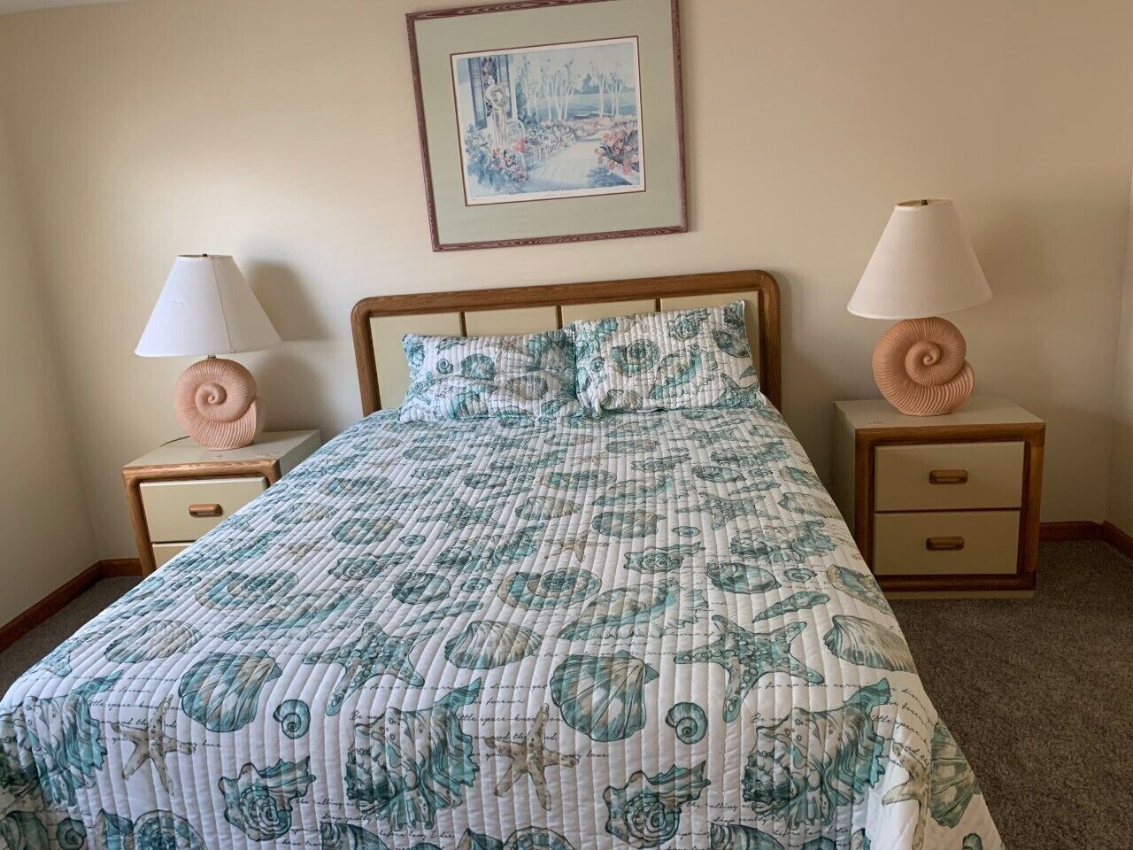 Une chambre à coucher avec un lit, deux tables de chevet avec des lampes, un tableau encadré au mur et un sol recouvert d’une moquette.