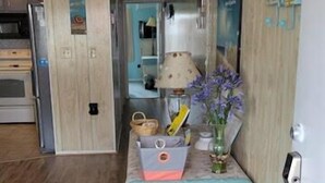 Un pasillo con piso de madera, ventilador de techo, una mesa con lámpara, un florero con flores y un letrero que dice 'Shoes Please'.