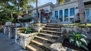 Une maison moderne avec un escalier en pierre menant à un espace extérieur couvert, entouré d’une végétation luxuriante et de palmiers.