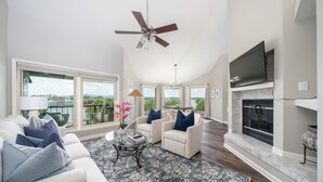 A spacious living room with a ceiling fan, a fireplace, a large TV, and a view of the outdoors. - Orchid On The Water - w/ Boat Slip On Lake Hamilton! (Hot Springs)