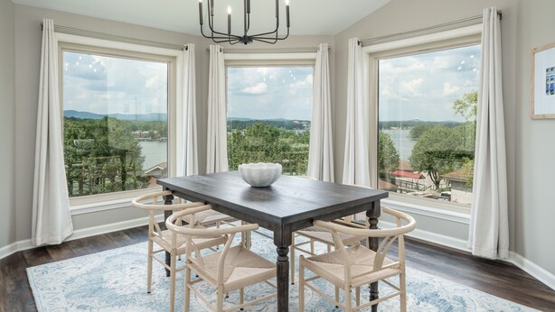 A dining area with a dark wooden table, four wicker chairs, and a chandelier. Three large windows offer views of a river and greenery. - Orchid On The Water - w/ Boat Slip On Lake Hamilton! (Hot Springs)