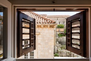 On aperçoit, à travers des volets en bois ouverts, un bâtiment en pierre avec un toit en tuiles, une ruelle étroite et un petit jardin.