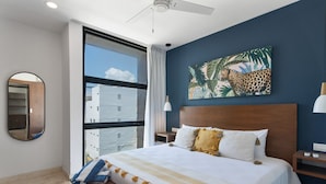 Une chambre d’hôtel avec un grand lit, une vue sur les immeubles par la fenêtre, un ventilateur de plafond et un tableau représentant un léopard au mur.