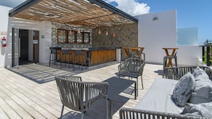 Un espace bar extérieur doté d’une pergola en bois, de sièges avec des coussins et d’un mur décoré de motifs.