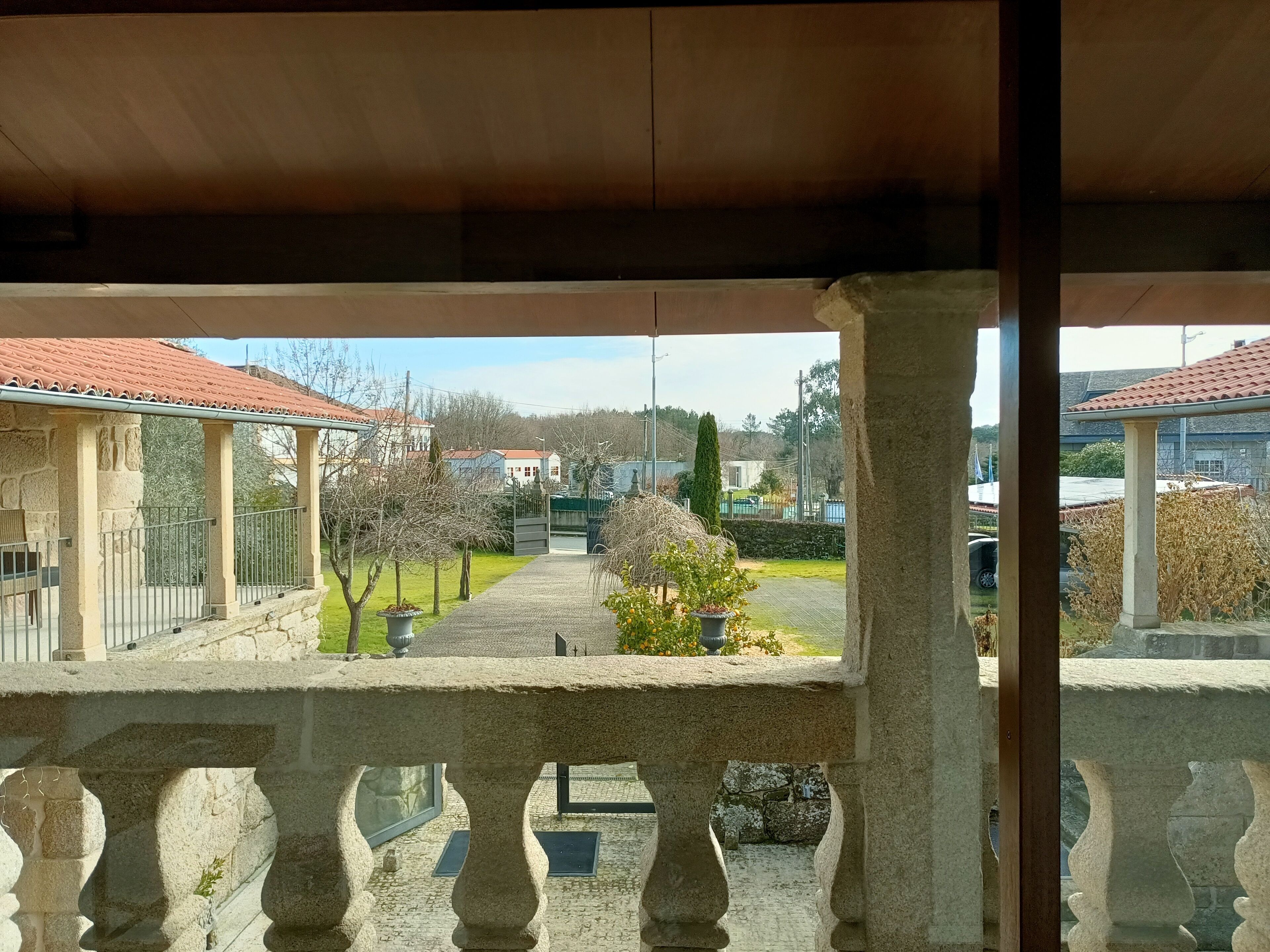 Een stenen balustrade met uitzicht op een binnenplaats, met een geplaveid pad, plantenpotten en gebouwen met rode pannendaken.