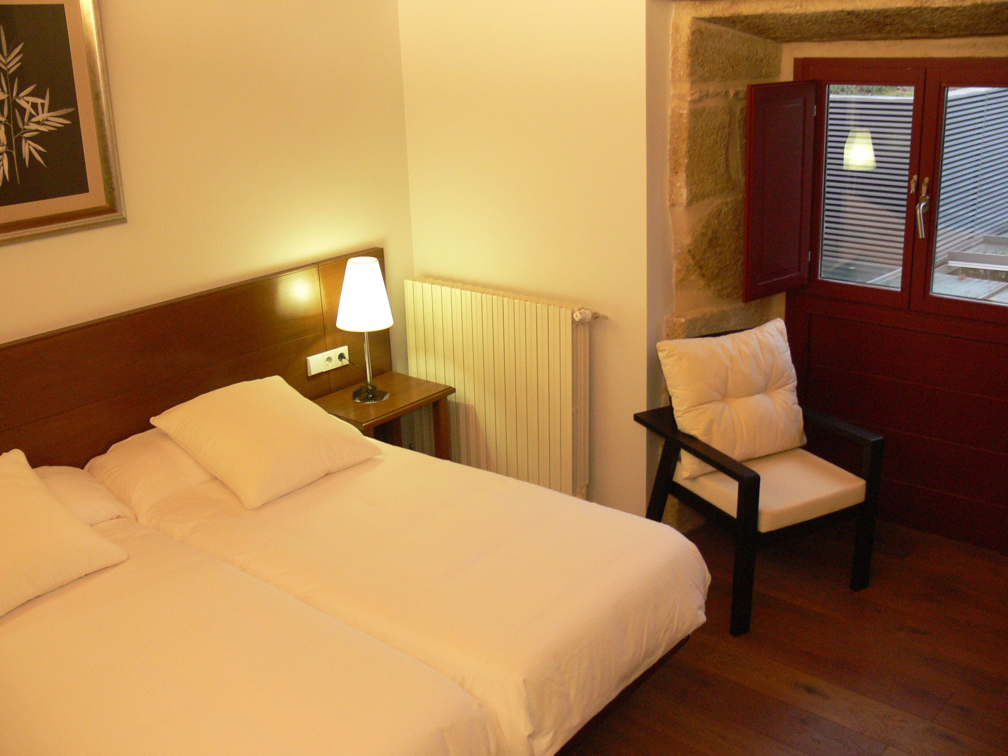 A hotel room with a double bed, a nightstand with a lamp, a chair, and a window with blinds.