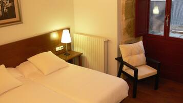 A hotel room with a double bed, a nightstand with a lamp, a chair, and a window with blinds.