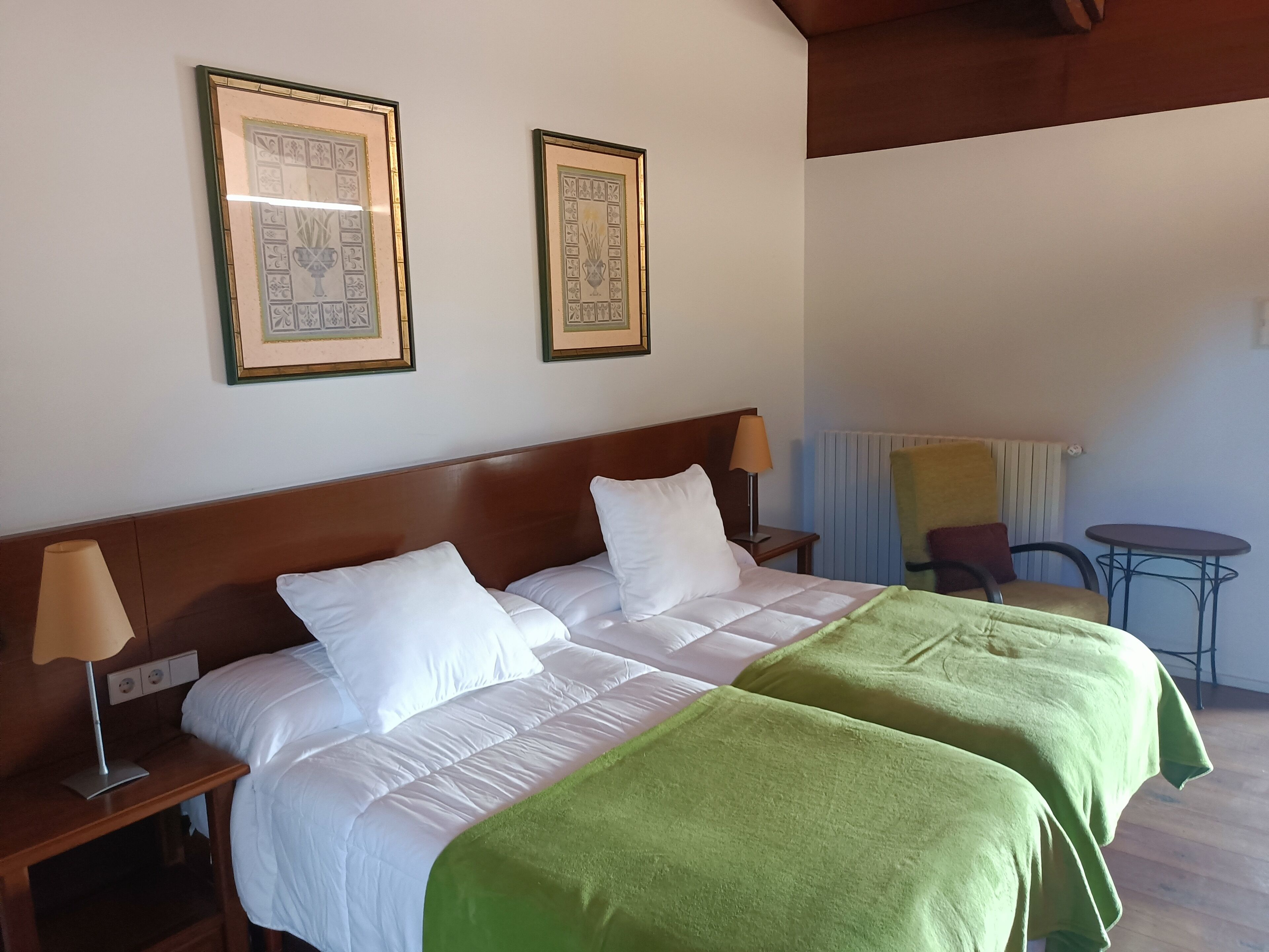 A double bed with white bedding and a green throw, a nightstand with a lamp, and two framed pictures on the wall.