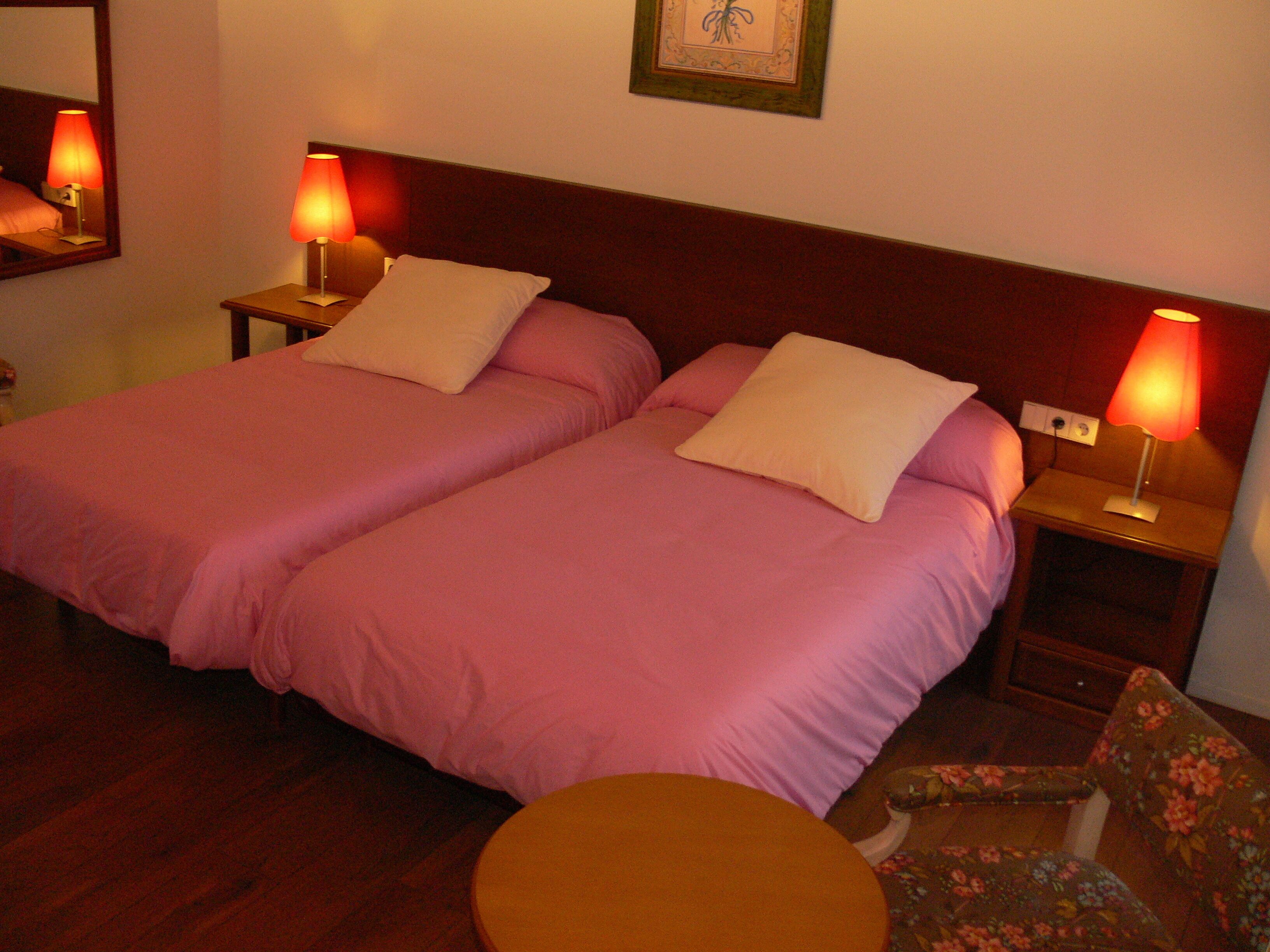 A hotel room with two beds, each with a pillow and a nightstand with a lamp. There is a chair and a small round table in the room.