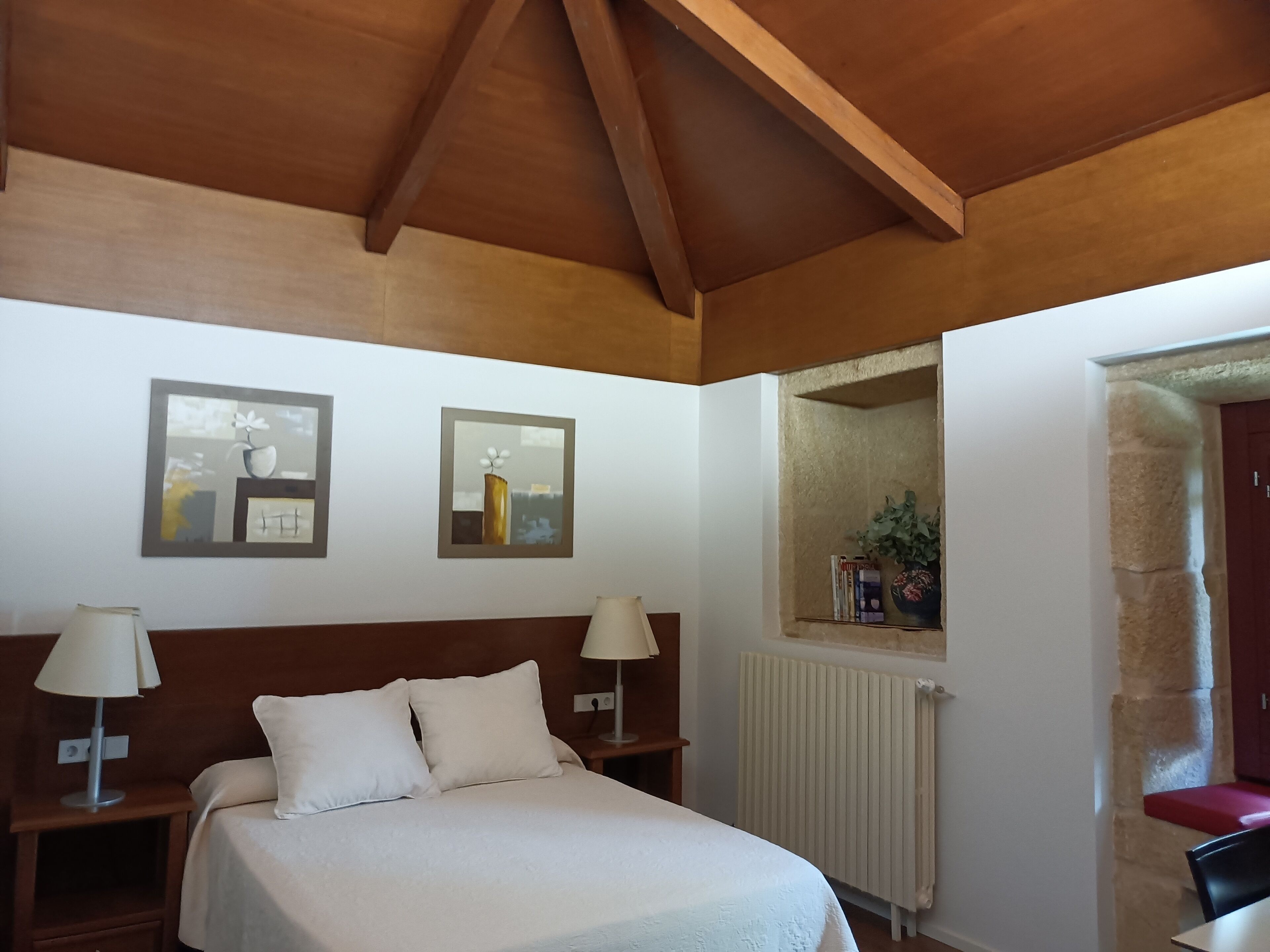 A bedroom with a wooden ceiling, a bed with white bedding, two bedside tables with lamps, and two framed artworks on the wall.
