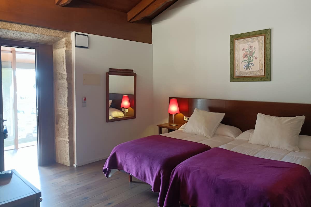 A hotel room with two beds, a wooden ceiling, a mirror, a nightstand with a lamp, and a framed picture on the wall.