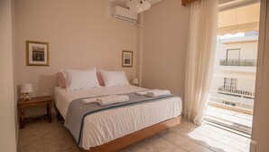A neatly made bed with white and pink pillows, a bedside table with a lamp, and a window with a view of a building. - Seafront Apartment Grace (Kalamata)