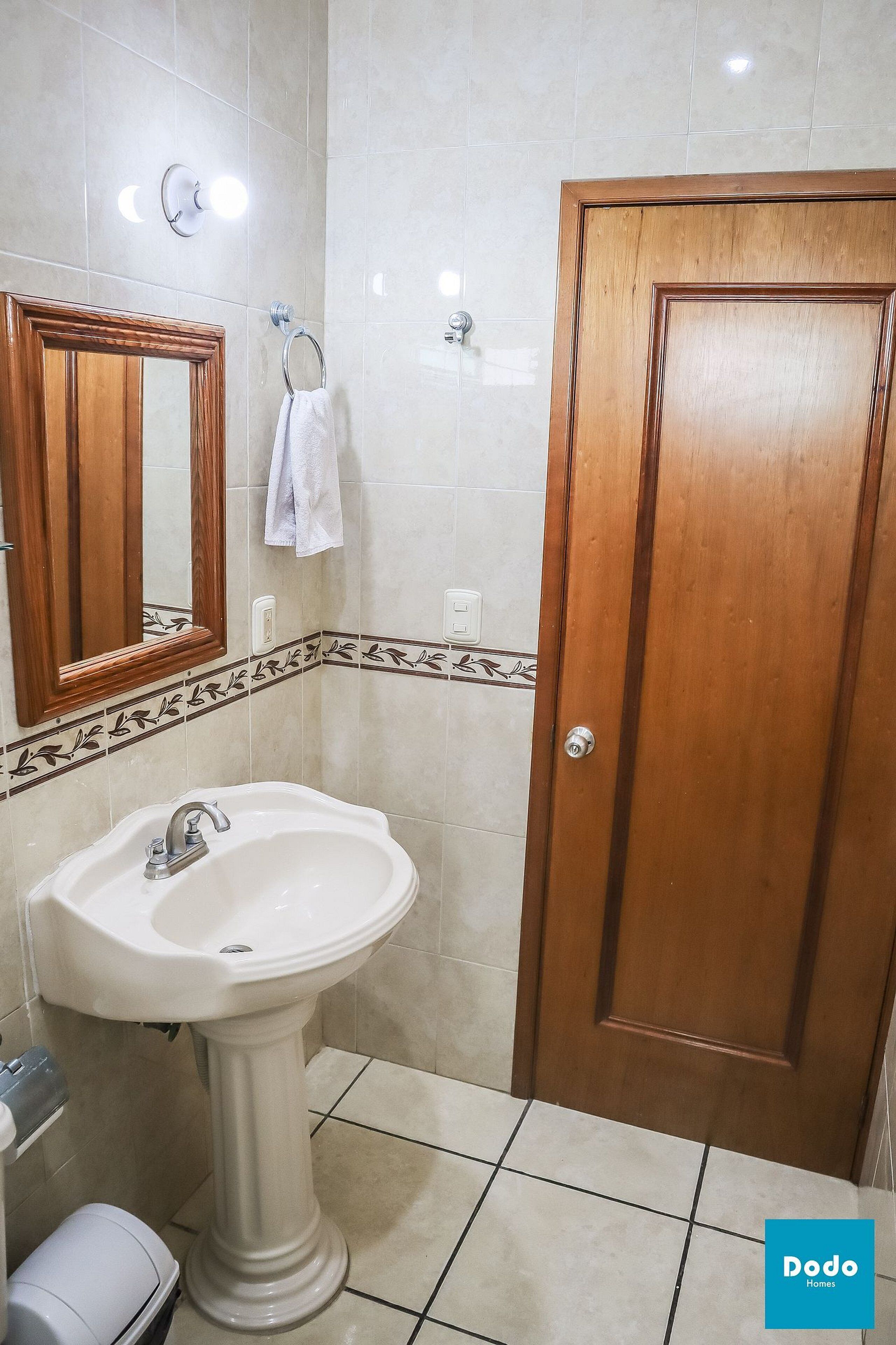 A bathroom with a pedestal sink, a wooden door, a mirror with a wooden frame, and a towel hanging on a metal ring.