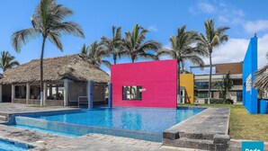Un área de piscina con una estructura de techo de paja, un edificio rosa brillante y palmeras.