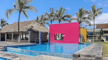 Un área de piscina con una estructura de techo de paja, un edificio rosa brillante y palmeras.