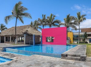 A swimming pool area with a thatched-roof structure, a bright pink building, and palm trees. - Cerritos Resort 138 (Mazatlán)