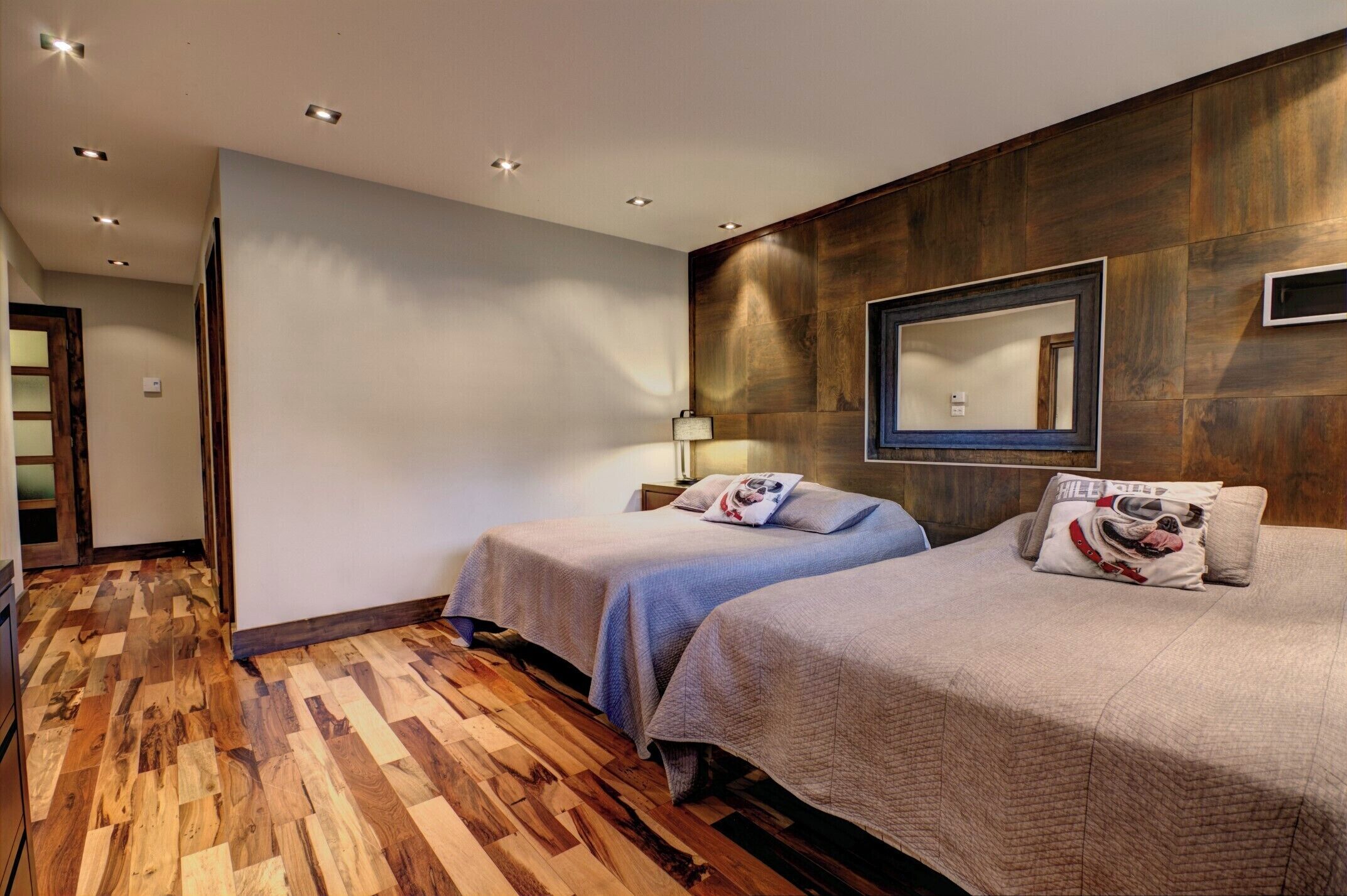 A hotel room with two beds, wooden flooring, a mirror on the wall, and a lamp.